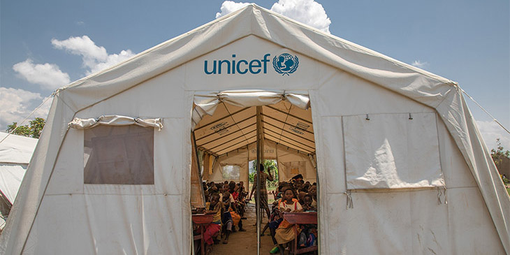 white tent with the unicef logo above the door from an african refugee camp. Inside a teacher is running a lesson with primary school children sitting at desks in rows