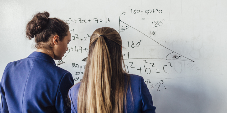 Two secondary school students in uniform facing a whiteboard with algebra equations written on it