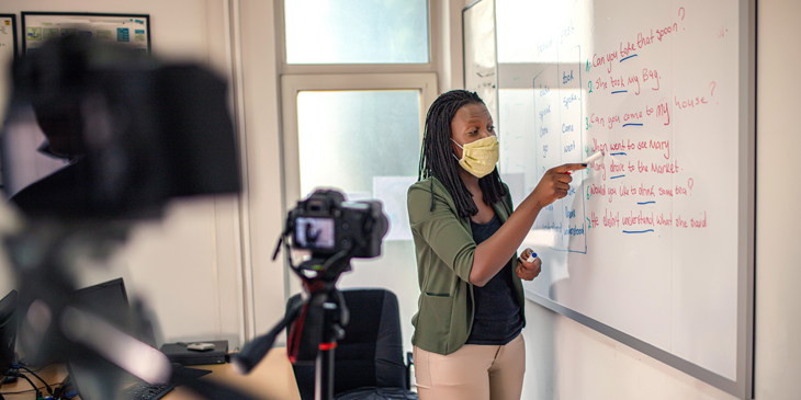 a teacher wearing a facemask being filmed for a virtual lesson as they write on a white board