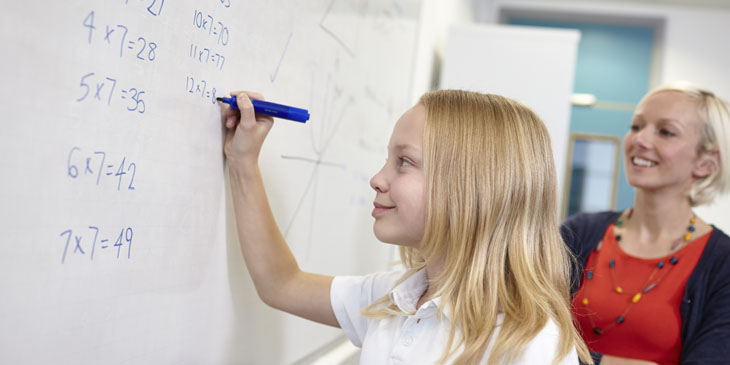 student writing sums on a white board closely observed by their teacher