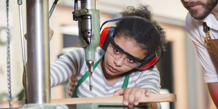 Woodwork student being observed by a teacher while working at a machine, wearing protective goggles and ear defenders