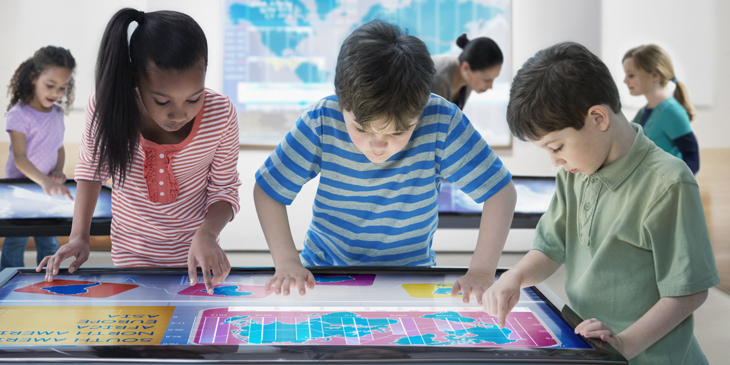 Three young learners looking at a map on a digital table top touch-screen display