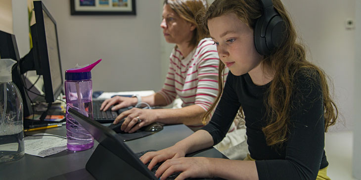 Secondary school student studying at home using a tablet and headphones with a parent working online in the background 