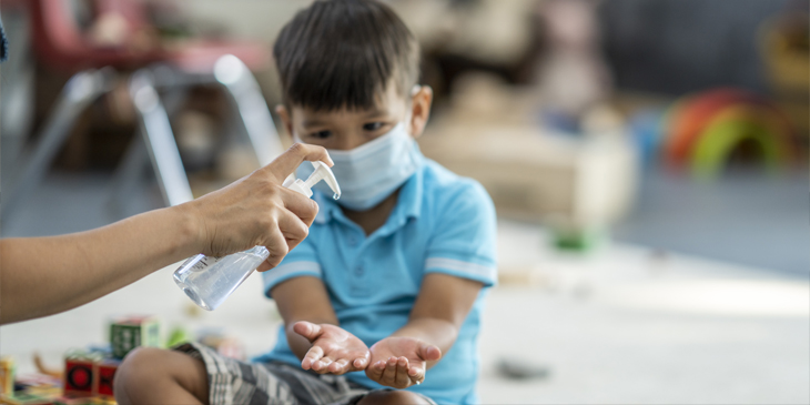 Primary school child sitting on the floor of a classroom wearing a facemask and having sanitiser put on their hands 